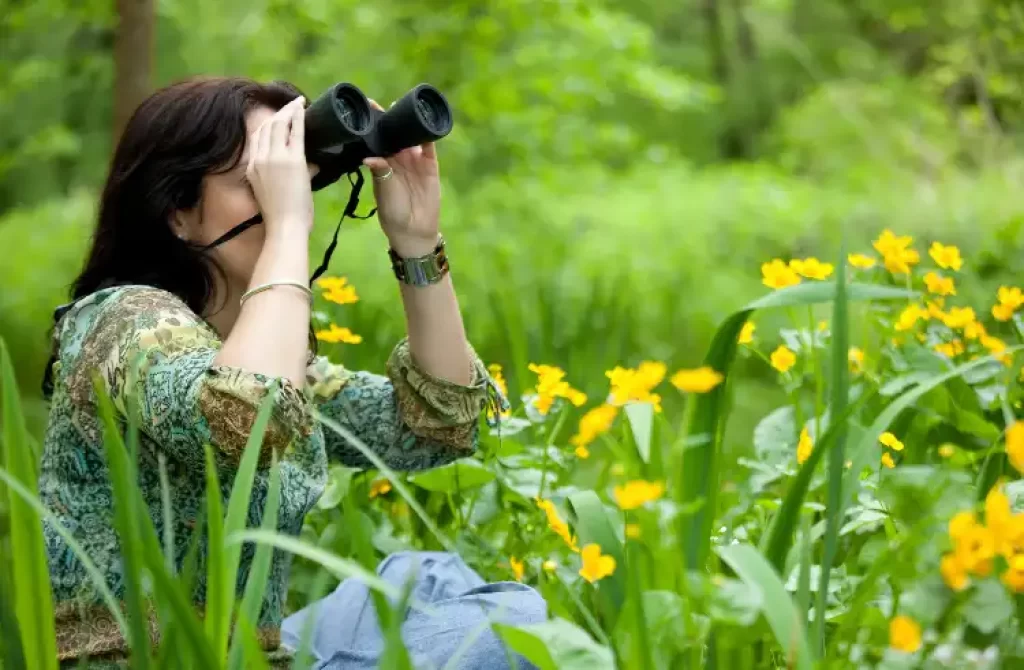 Woman in green floral blouse and jeans birdwatching with binoculars in a field of yellow flowers.